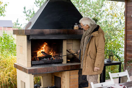 Contemporary senior man in warm casualwear standing by fireplace by houseの写真素材