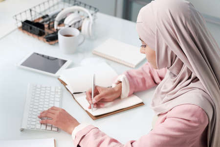 Contemporary young Muslim woman in hijab and smart casualwear making notesの写真素材
