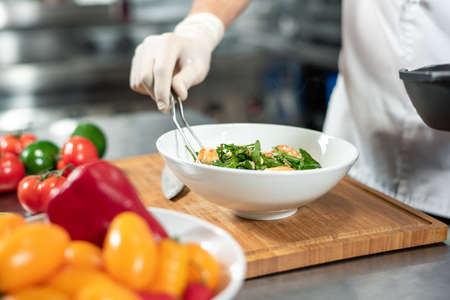 Gloved hand of young chef with fork putting cooked meal into white ceramic bowlの写真素材