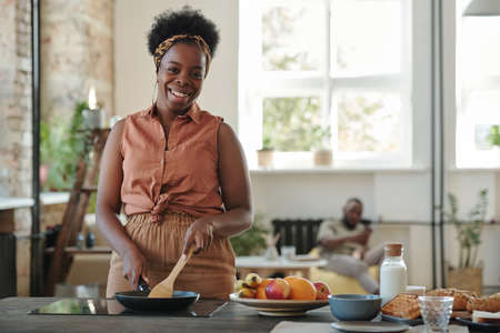 Young happy woman of African ethnicity cooking dinner for her family by stoveの写真素材