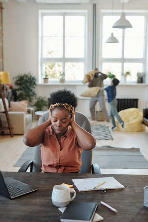 Young annoyed African woman with headache touching head while sitting by tableの写真素材