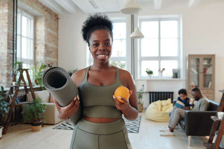 Healthy young African fit woman with toothy smile holding rolled mat and orangeの写真素材