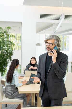 Confident mature businessman in eyeglasses talking to client on mobile phone