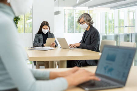 Two busy colleagues in smart casualwear and protective masks networking by tableの写真素材