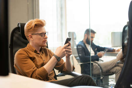 Young serious businessman in eyeglasses texting in smartphone in armchairの写真素材