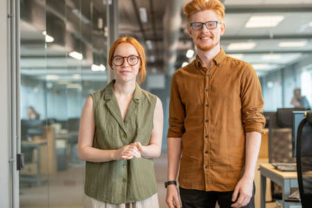 Young cheerful red haired colleagues in smart casualwear standing by each otherの写真素材