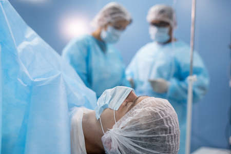 Young patient in mask and headwear lying on operation table against surgeons