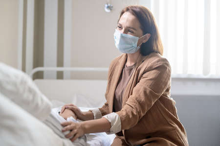 Young woman in casualwear and protective mask touching hands of sick friendの写真素材