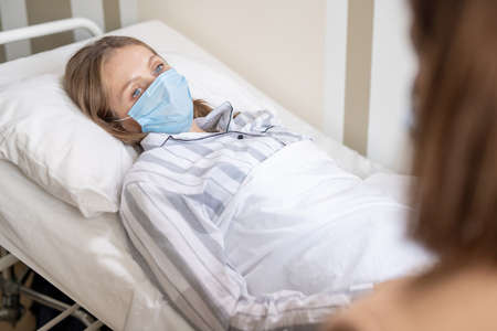 Sick female in pajamas and protective mask lying in bed in hospital chamberの写真素材