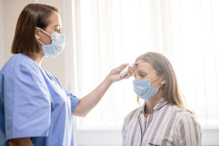 Young nurse in blue uniform and mask holding thermometer by patient forehead
