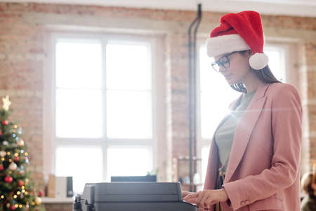 Young female office worker in xmas cap and eyeglasses working by xerox machineの写真素材