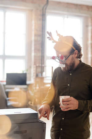 Young businessman in xmas eyeglasses pressing start button to copy document