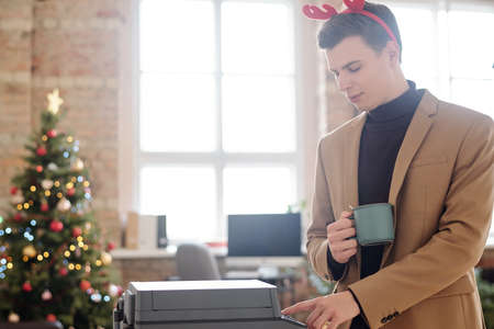 Young elegant businessman in xmas headband pushing button of xerox machine