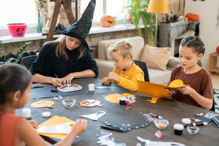 Concentrated young art teacher in witches hat sitting at table and drawing Halloween pictures with kids while preparing paper decorationsの写真素材