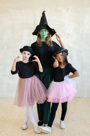 Portrait of serious green-faced woman in witches hat posing with cute mime girls grimacing against...