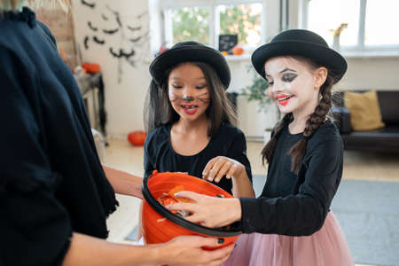 Cheerful beautiful multi-ethnic girls in Halloween costumes taking sweets from bucket held by woman, trick-or-treating tradition on Halloween conceptの写真素材
