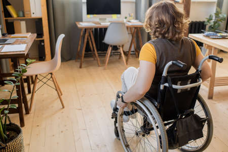 Back view of young brunette disable female in wheelchair moving towards table