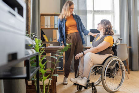 Young blond woman in casualwear looking at her disable friend in wheelchairの写真素材