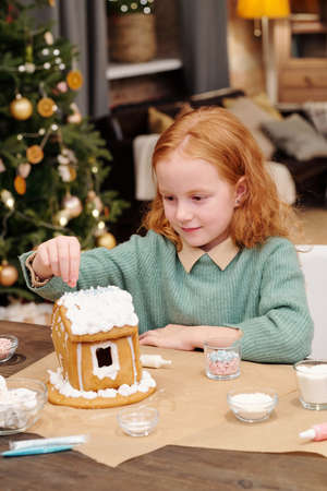 Cute girl sprinkling roof of gingerbread house decorated with whipped cream