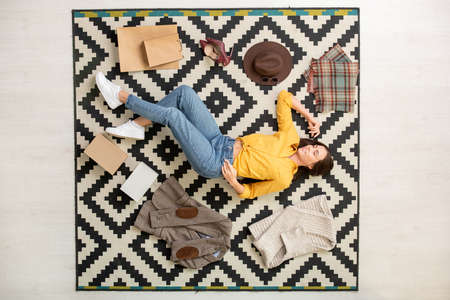 View of young happy woman surrounded by new smart casualwear lying on the carpetの写真素材