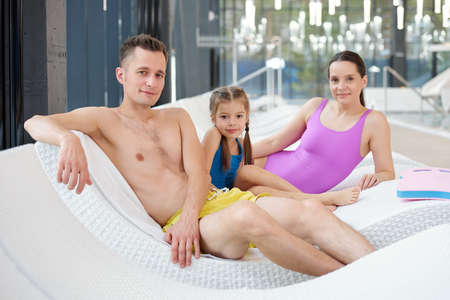 Young contemporary family relaxing on deckchairs by poolside at leisure center