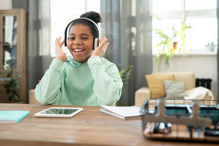 Cute cheerful schoolgirl of African ethnicity in headphones listening to music