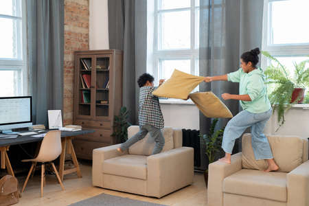 Two cute mixed-race siblings in casualwear pillow fighting on soft armchairs