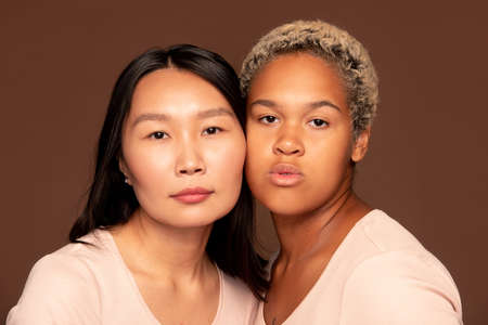 Two young intercultural females looking at camera while touching by facesの写真素材