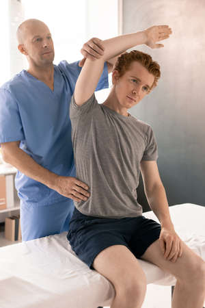 Bald mature physiotherapist in uniform supporting arm of exercising patient