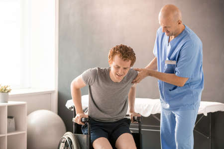 Mature clinician in uniform helping young man in pain to sit in wheelchairの写真素材