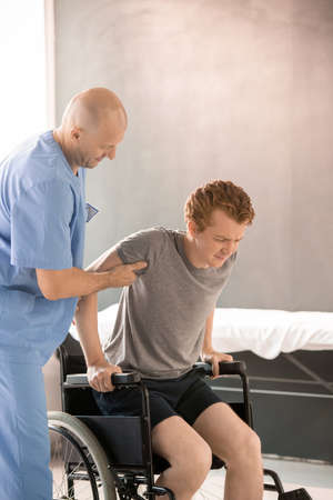 Bald physiotherapist helping sick young man to sit in wheelchair after trainingの写真素材