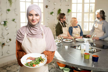Happy young Muslim female in hijab holding plate with selfmade vegetable saladの写真素材