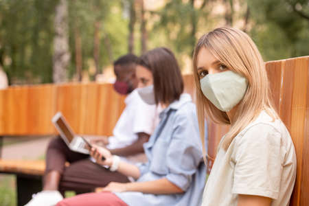 Young blond woman in protective mask looking at you against two millennialsの写真素材