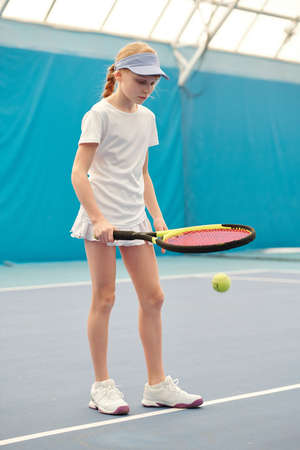 Youthful blond girl in white activewear pushing ball by tennis racket on stadiumの写真素材