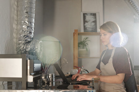 Serious young craftswoman in apron networking by table in workshop or studio