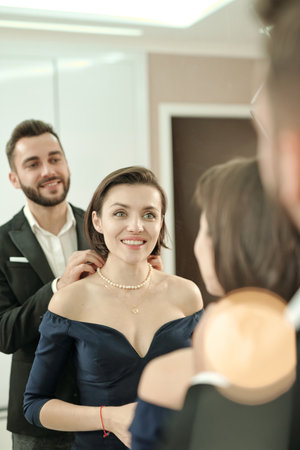 Happy young beautiful woman in black evening gown standing in front of mirror