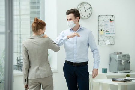 Two young office managers in protective masks giving each other elbow bumpの写真素材