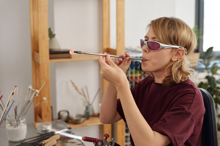 Young skilled female lampworking master blowing into long tube by workplaceの写真素材