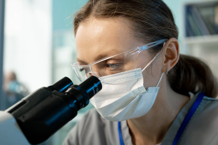 Young female scientist looking at chemical or biological samples in microscope