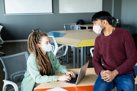 Two young intercultural office workers in protective masks looking at each otherの写真素材