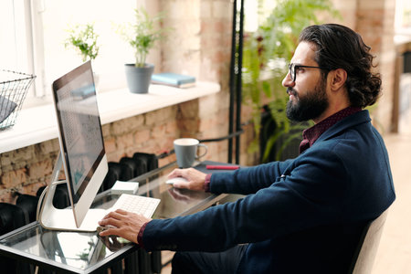 Serious young bearded broker or economist in suit sitting in front of computerの写真素材