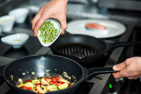 Hand of chef putting frozen green peas into frying pan with chopped vegetablesの写真素材