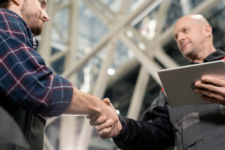 Mature foreman with digital tablet greeting his young subordinate by handshake