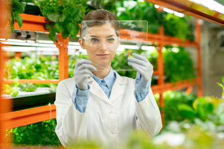 Young female worker of vertical farm holding square glass in front of her faceの写真素材