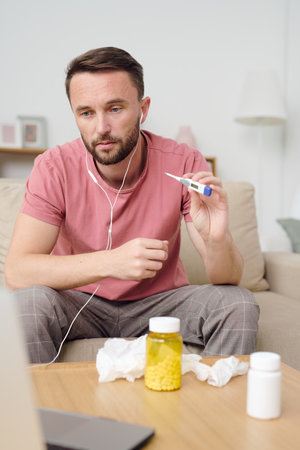 Young sick male patient showing thermometer with his body temperature to doctor