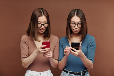 Young Asian twin sisters in casualwear standing close to one another and textingの写真素材