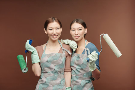 Happy young Asian female twins in aprons and gloves holding paint rollersの写真素材