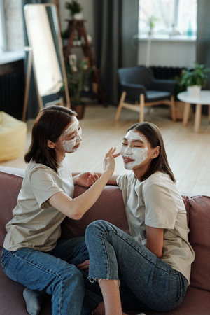 Young brunette twins with facial clay mask sitting on soft couch in living-roomの写真素材