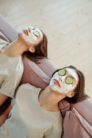 Young female twins with clay mask on faces and fresh cucumber slices on eyesの写真素材