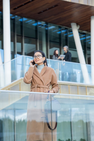 Young Asian businesswoman in elegant beige coat talking on mobile phoneの写真素材